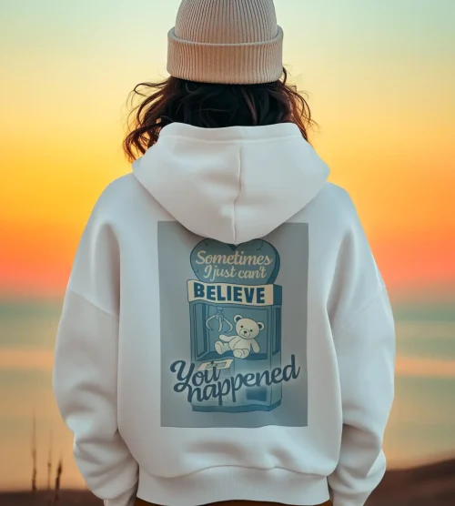 A woman wearing a beige knit beanie and white hoodie with a teddy bear graphic, standing on the beach during sunset.
