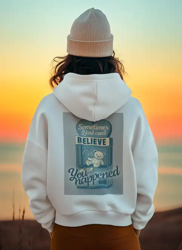 A woman wearing a beige knit beanie and white hoodie with a teddy bear graphic, standing on the beach during sunset.