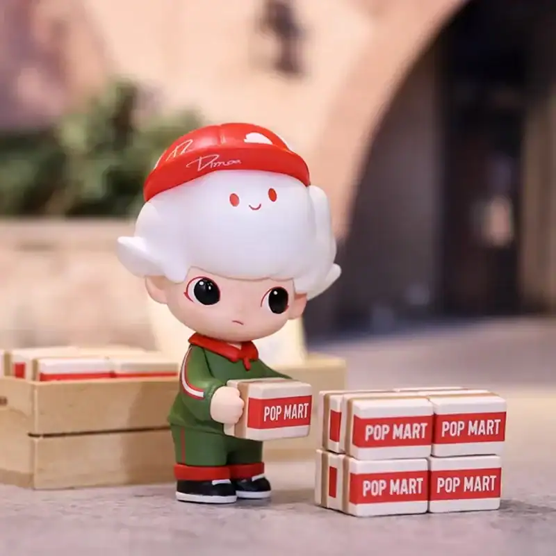 Cute cartoon-style figurine with oversized head and expressive eyes, holding a small box labeled "POP MART," sitting among similar boxes, in a cozy indoor setting.